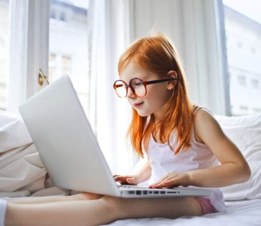 Rudowłose dziecko w okularach korzysta z laptopa na białym łóżku